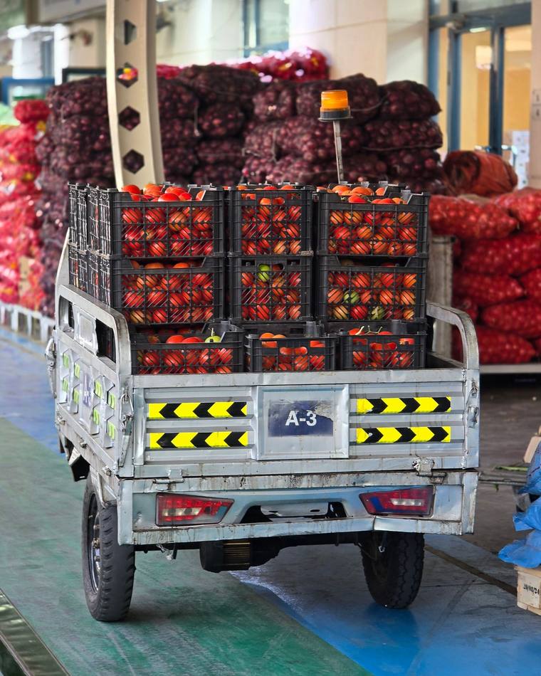 A loader stacked with crates of tomatoes on a wholesale market floor, sacks of onions stacked behind it
